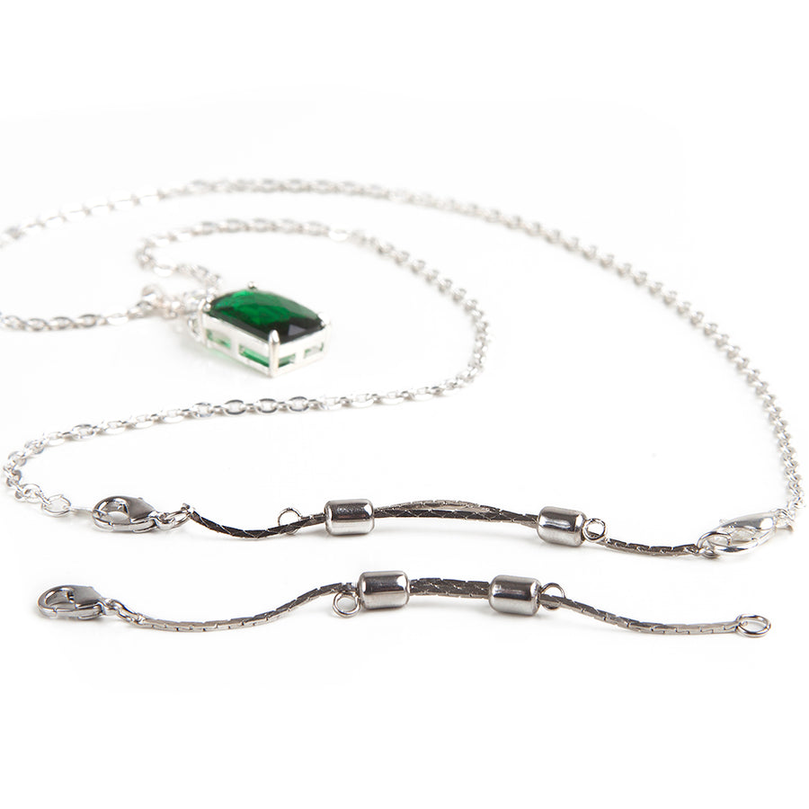 One silver adjustable necklace extender in front of an emerald and silver necklace with attached silver adjustable necklace extender.
